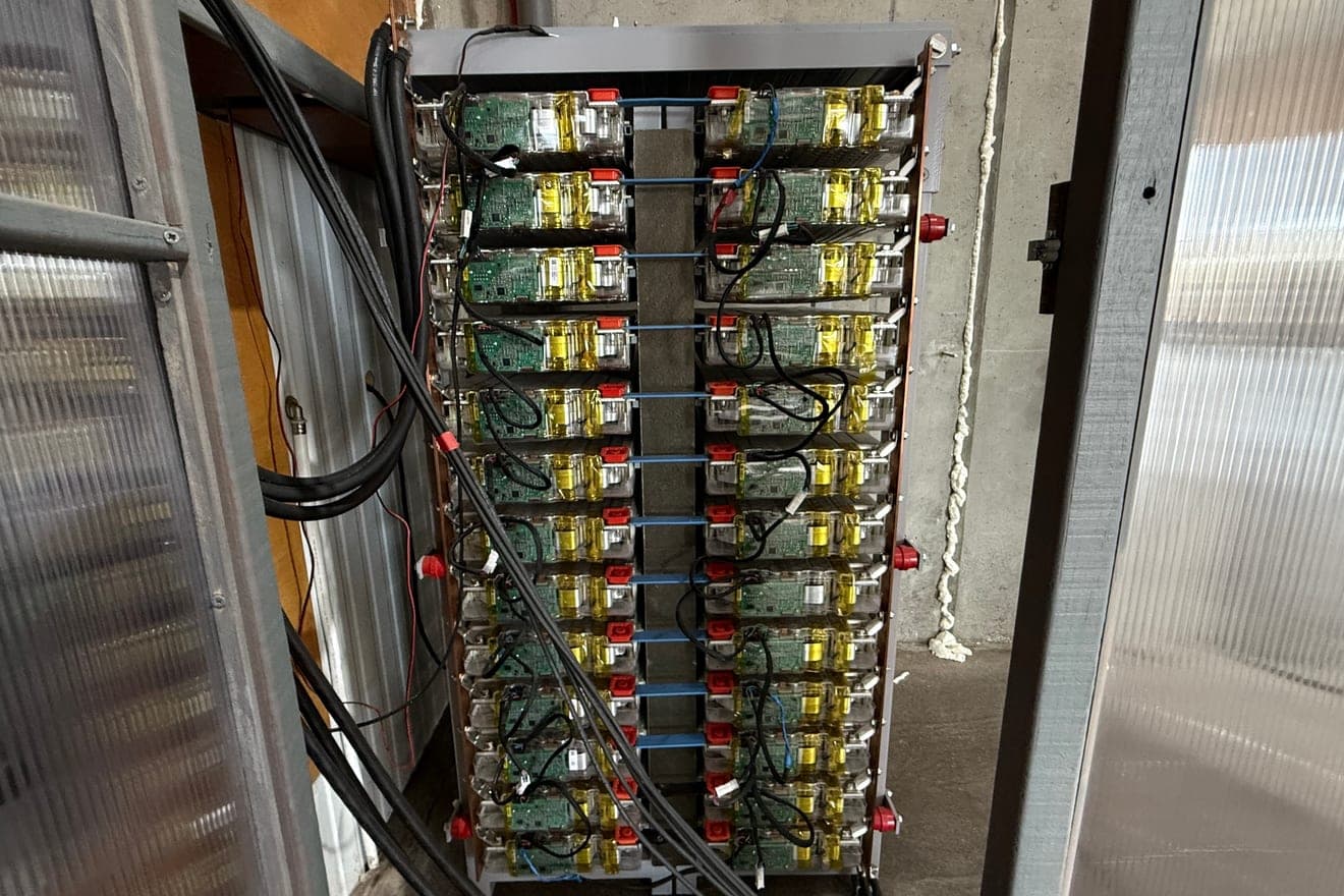 Rack de baterias entre duas paredes de policarbonato, com módulos empilhados em duas colunas, cada um com uma placa eletrónica verde à vista e ligações em cobre nas laterais.