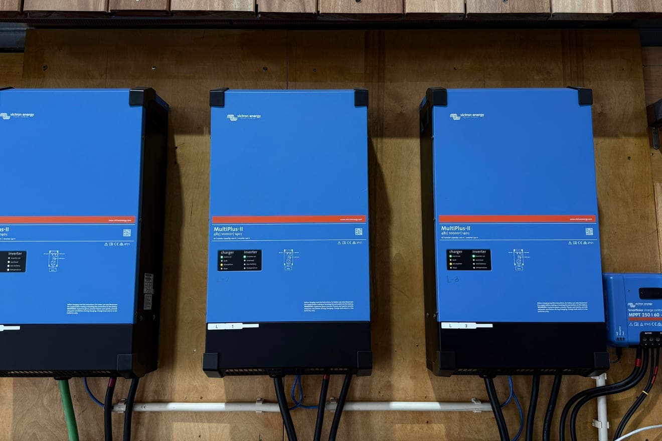 Três inversores Victron MultiPlus-II azuis montados lado a lado num painel de contraplacado, com um regulador de carga SmartSolar mais pequeno à direita e os cabos a descer para uma calha por baixo.